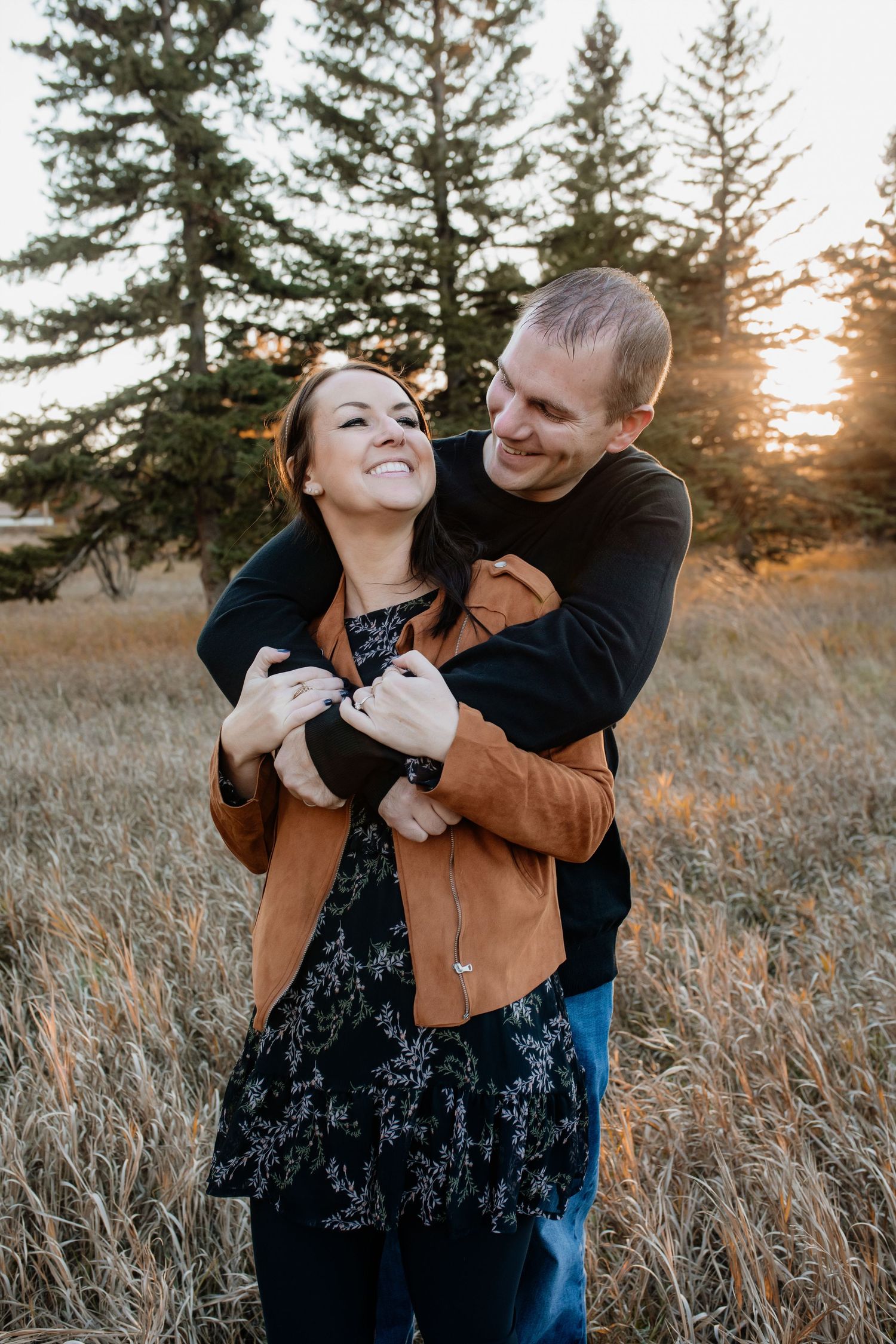 Happy couple embracing and laughing together in a golden field with pine trees glowing in sunset light.