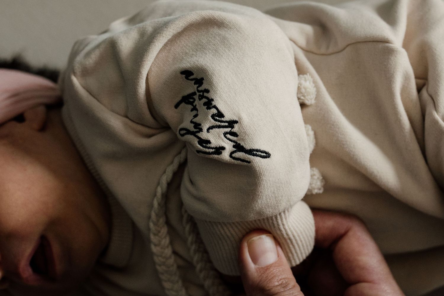 Close-up of newborn baby sleeping in a cream outfit with embroidered cursive name detail on the sleeve.
