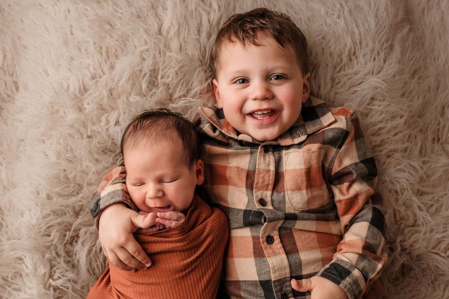Smiling toddler boy in plaid shirt lying beside sleeping newborn sibling on fur blanket.
