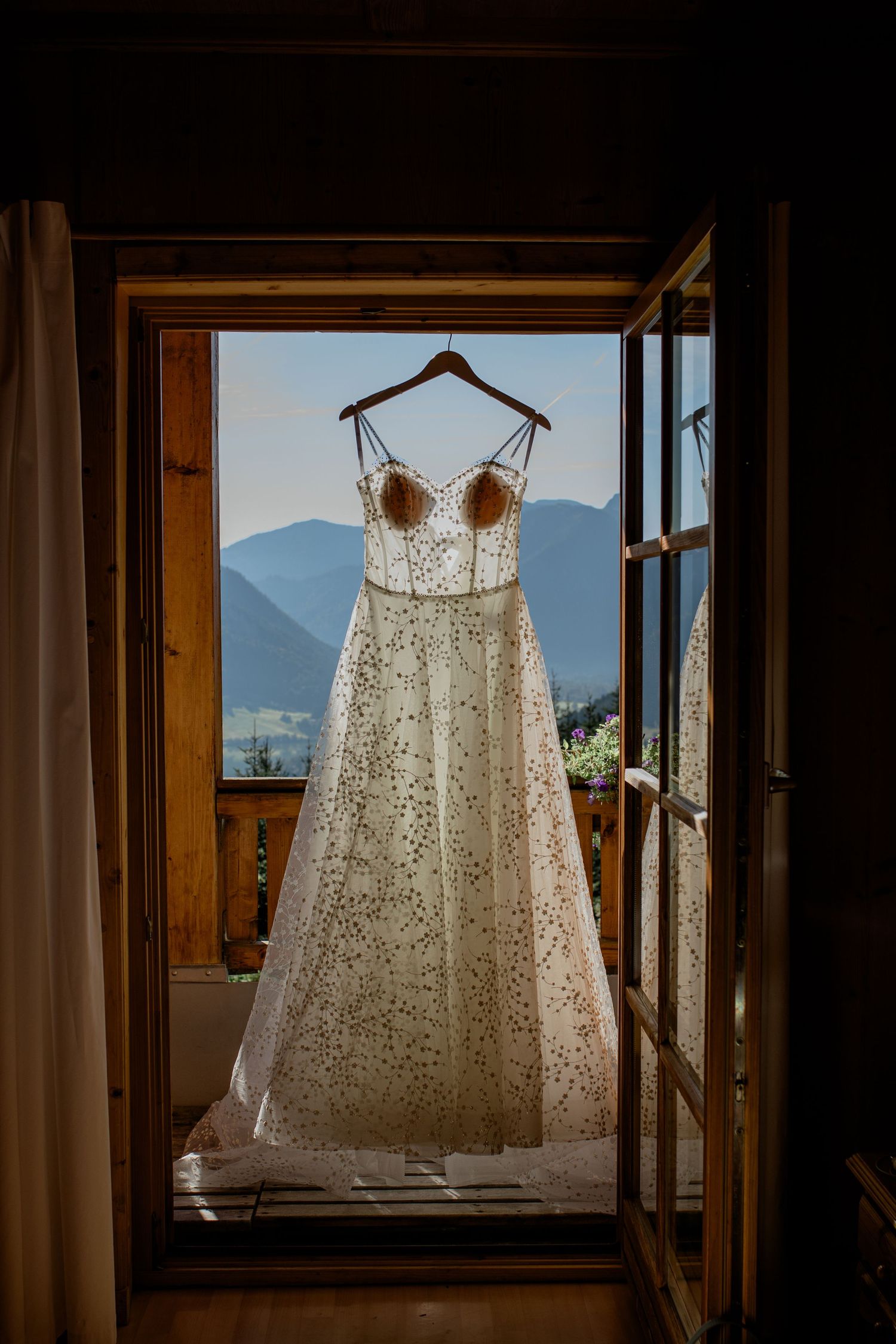 Elegant lace wedding dress hanging in a wooden window frame with stunning mountain views in the background.
