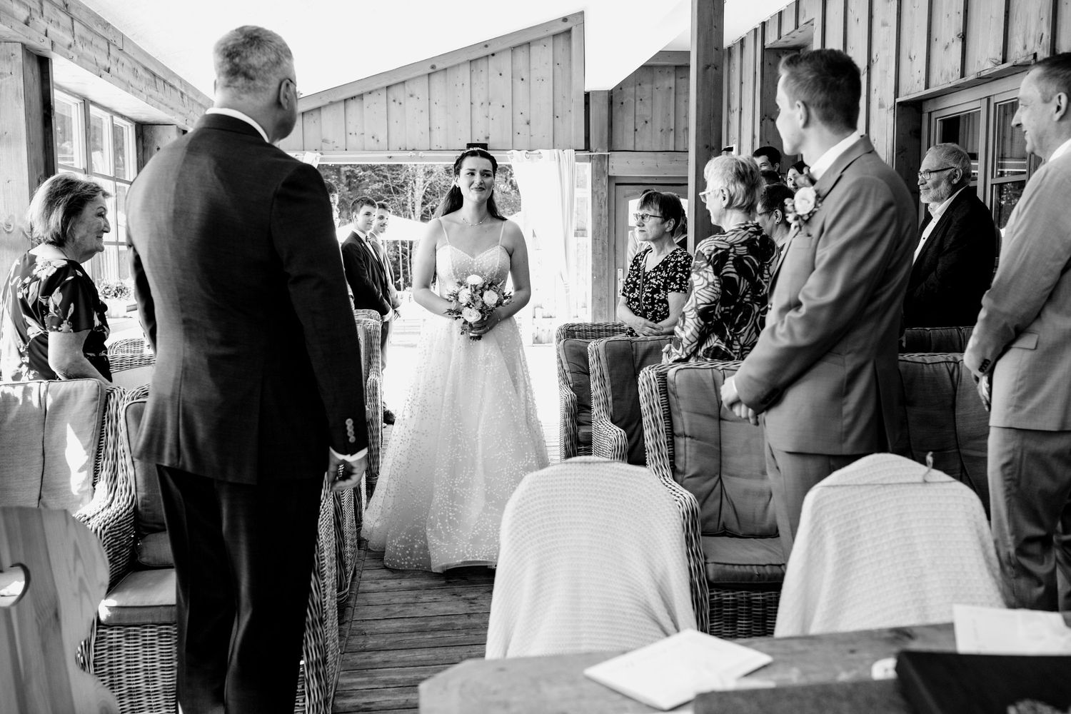 Black and white photo of a bride walking down the aisle at an outdoor wedding ceremony with seated guests.