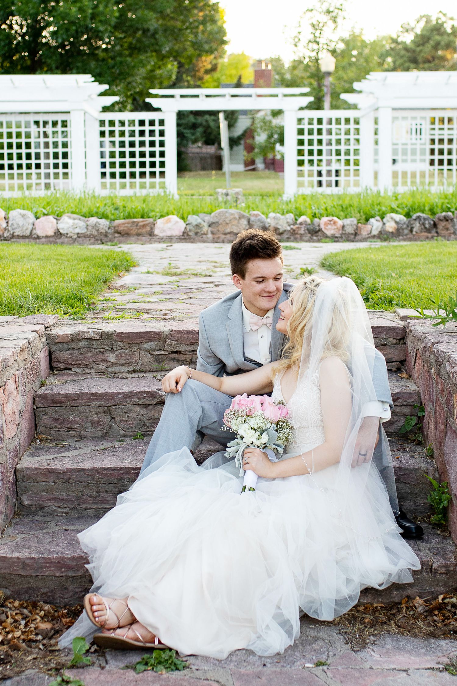 Bride and groom sitting on stone steps in a garden, sharing a romantic moment with a white floral bouquet.