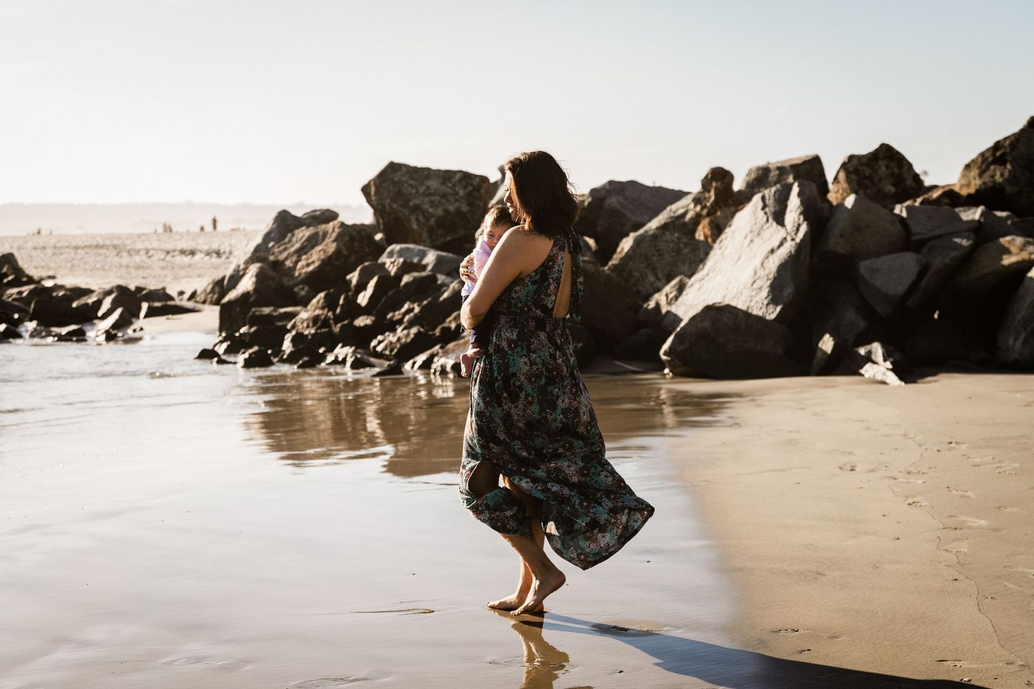 Woman in floral maxi dress walking barefoot along sandy beach with large rocks behind her.