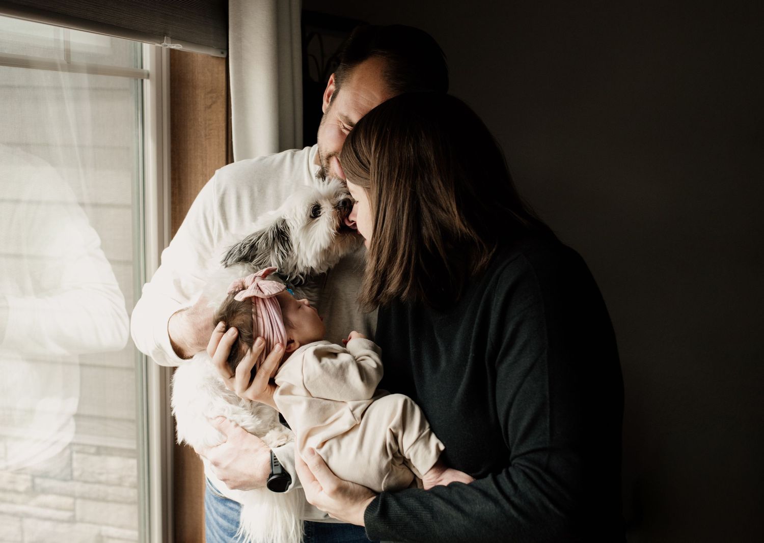 Couple standing by a window sharing a tender moment with their newborn baby and small white dog.