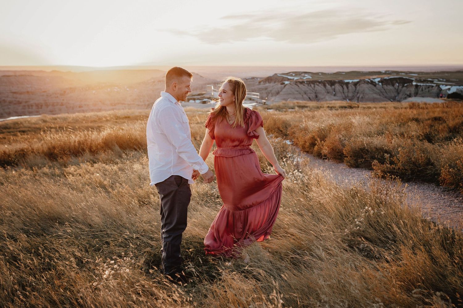Couple holding hands and dancing in a golden field at sunset with scenic landscape.