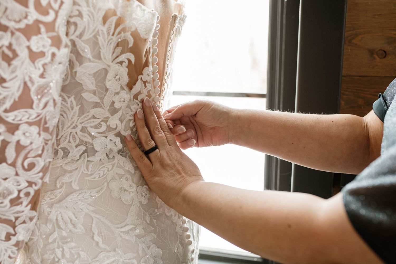 Person fastening buttons on an intricate lace wedding dress near a window.