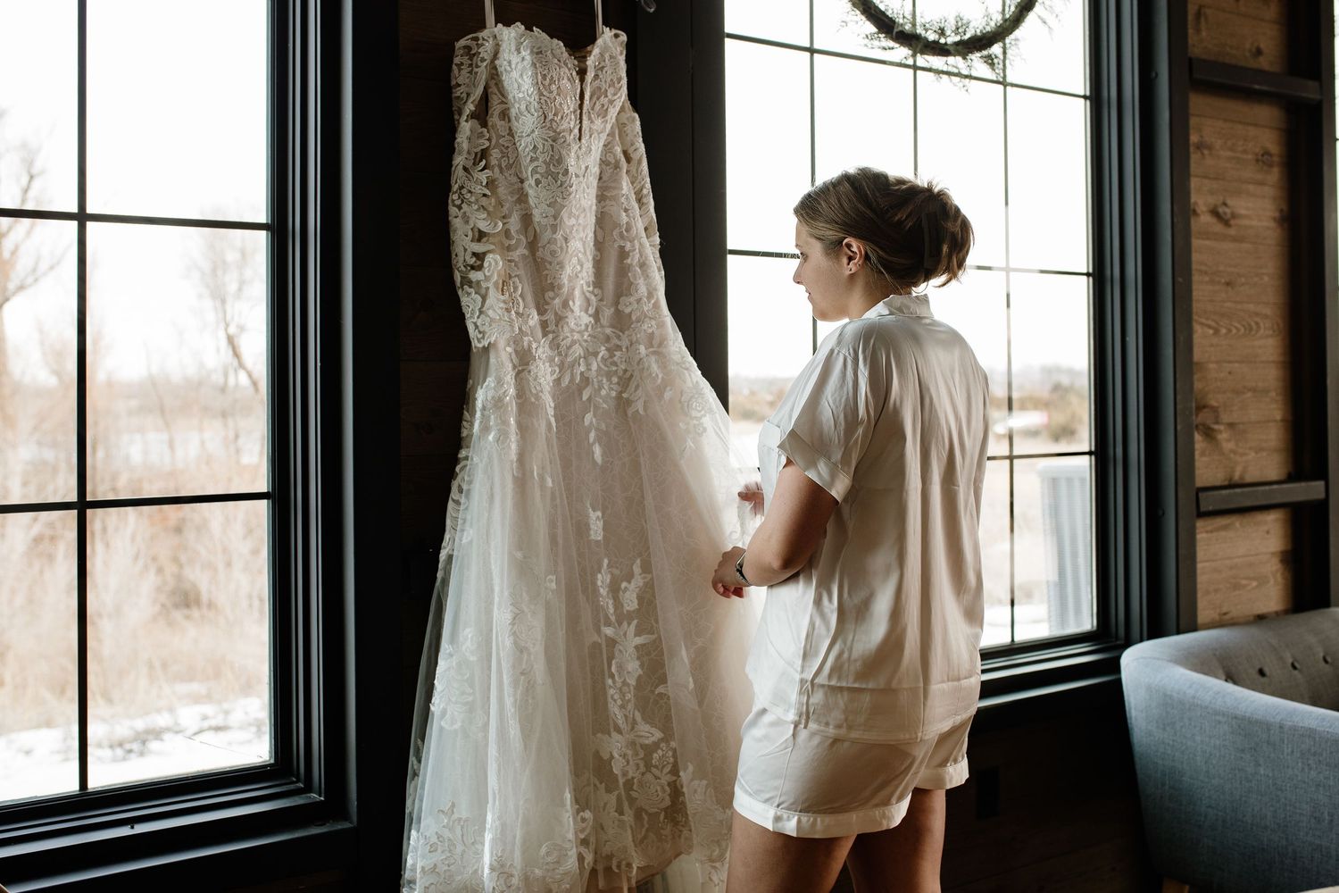 Bride in white robe admiring her lace wedding dress hanging by a large window in a rustic venue.