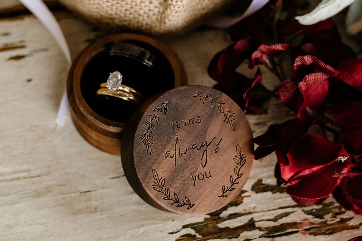 Wooden ring box engraved with 'it was always you' beside floral sprigs, holding two wedding rings on rustic surface.