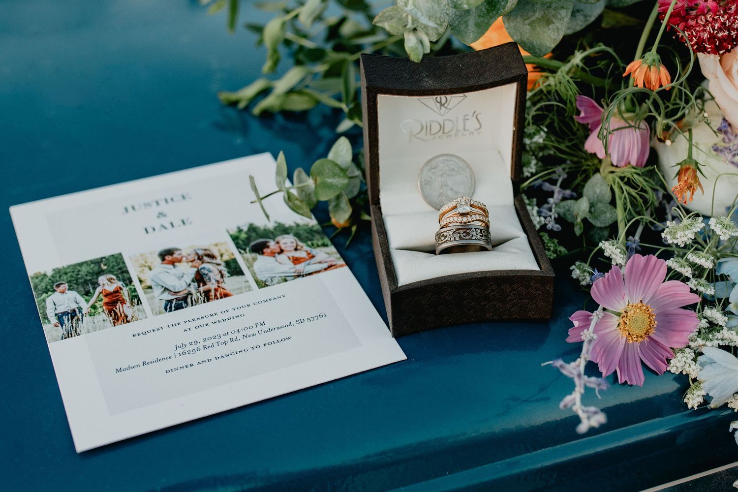 Wedding invitation, stacked rings in a jewelry box, and a colorful floral bouquet displayed on a blue surface.