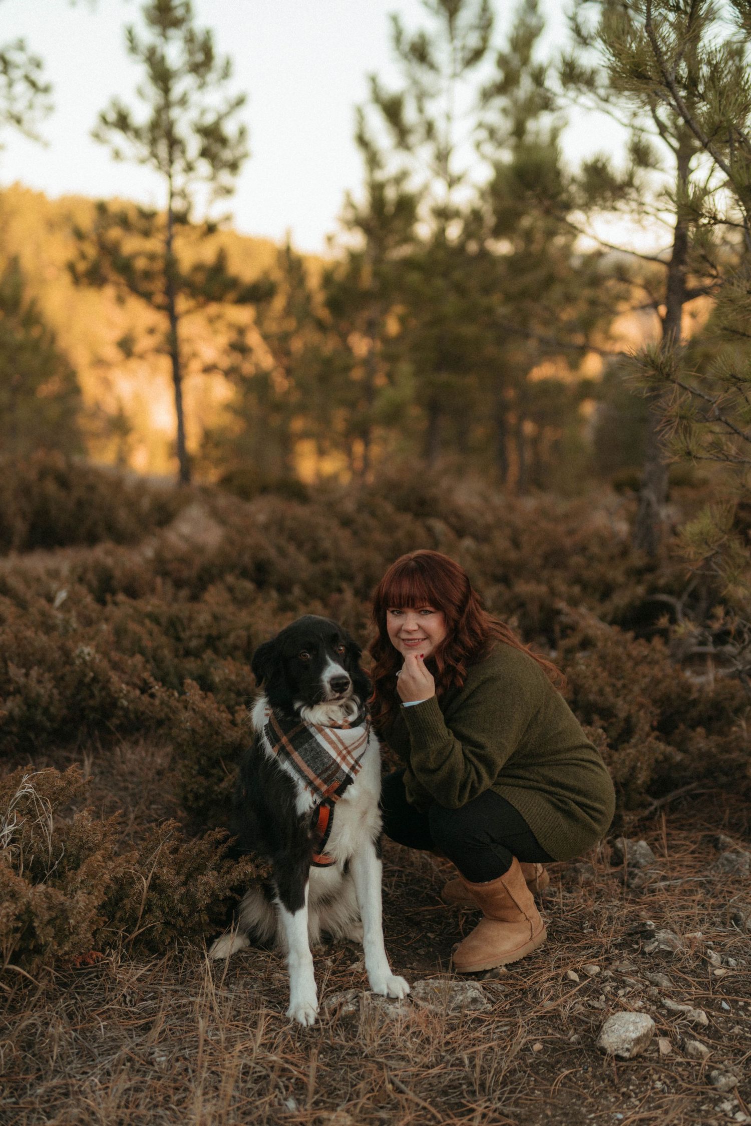 Woman with red hair crouching beside a black and white Border Collie wearing a plaid bandana in a forest setting.
