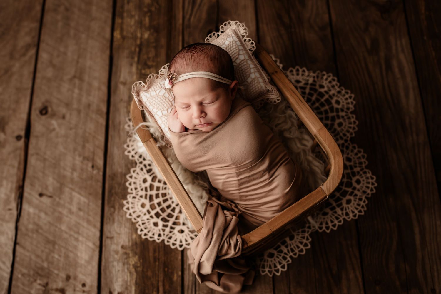 Sleeping newborn baby swaddled in beige wrap lying in wooden box on lace doily rustic floor.