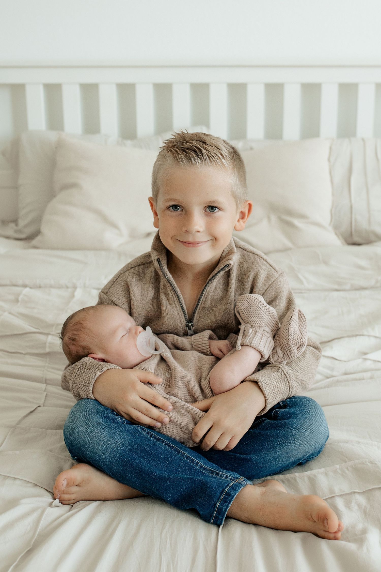 Smiling young boy sitting cross-legged on bed gently holding sleeping newborn baby sibling in his arms.