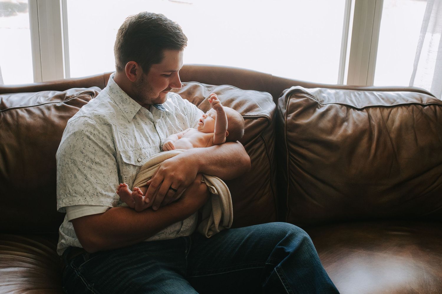 New father sitting on leather couch gazing lovingly at his sleeping newborn baby.
