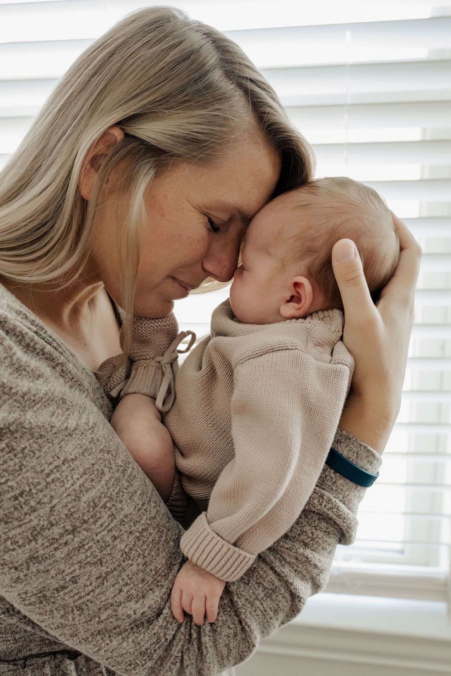A blonde mother gently presses her forehead against her sleeping newborn baby while holding them close indoors.