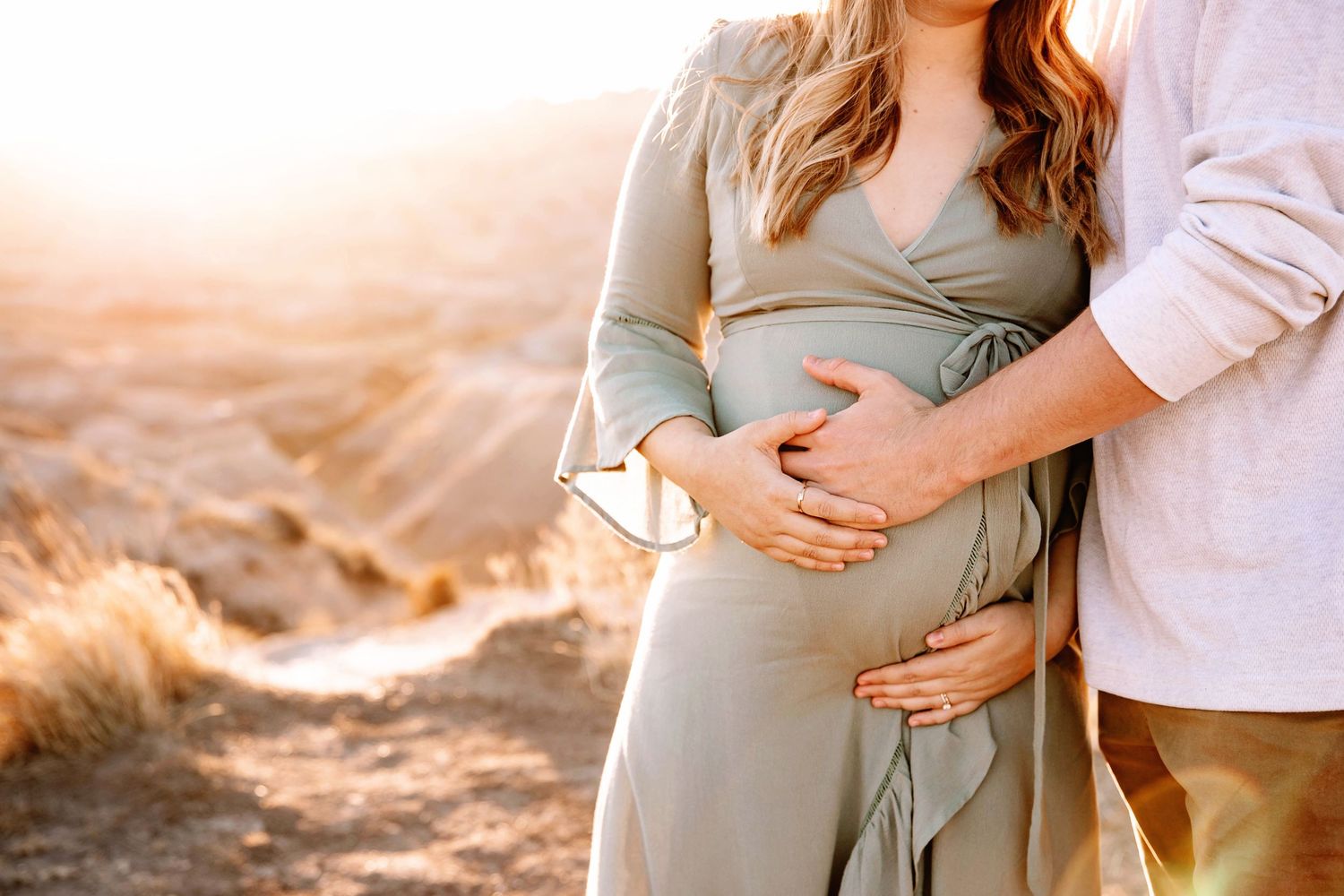 Partner embraces pregnant woman in sage wrap dress from behind in sunlit desert setting.