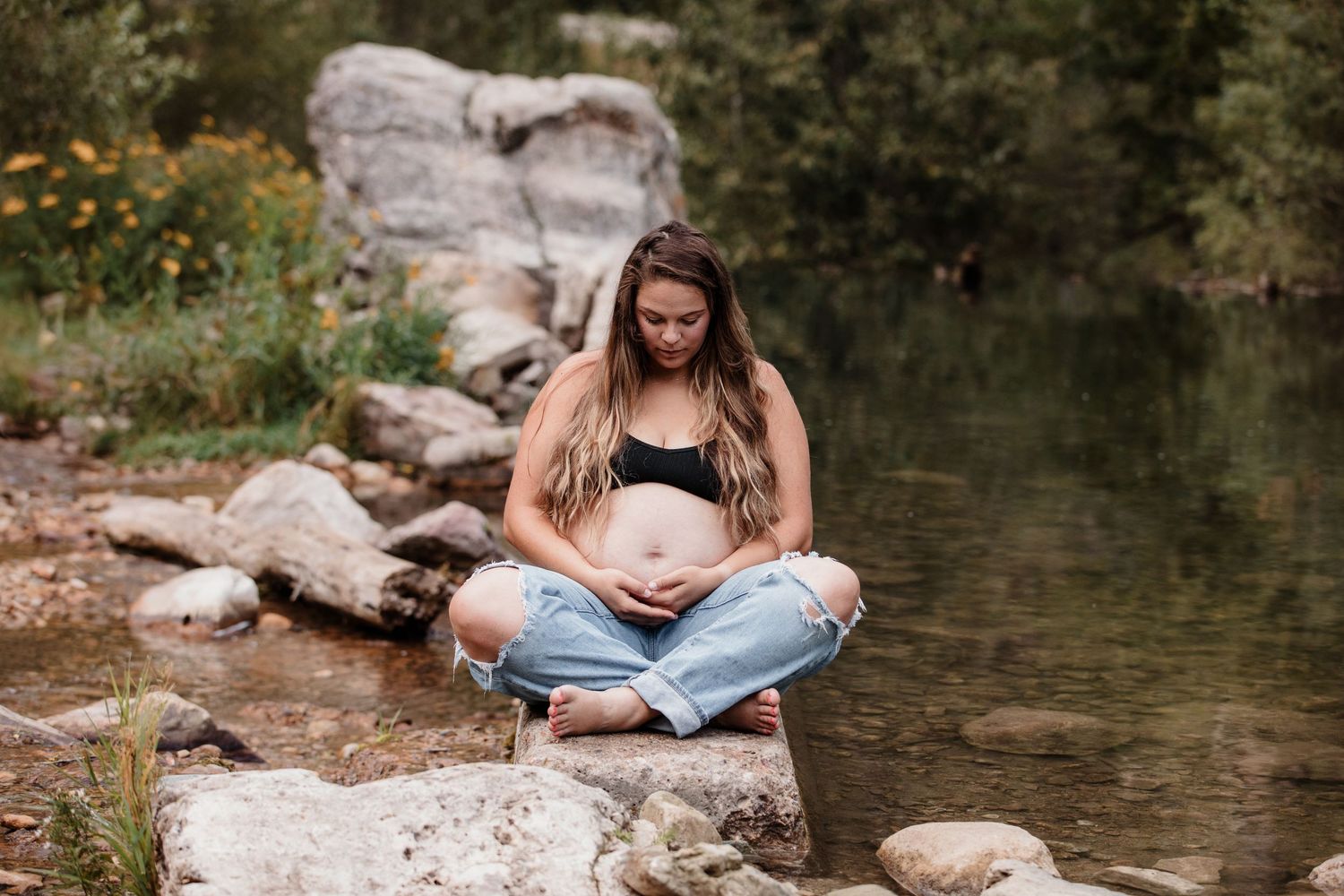 Pregnant woman in jeans sits cross-legged on rocks by a stream, cradling her baby bump.