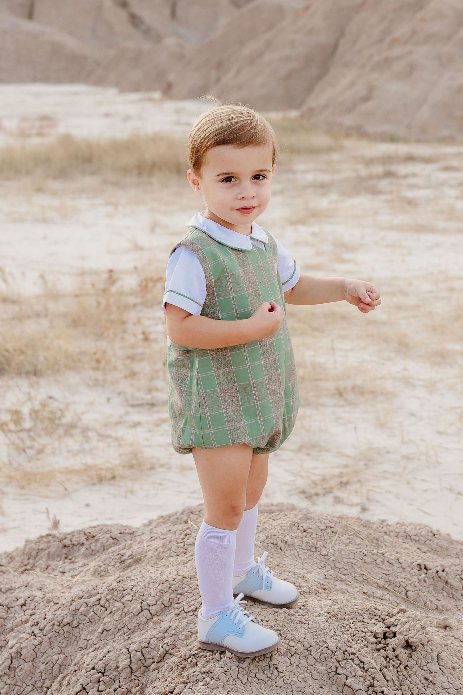 Adorable toddler boy in green plaid romper and white knee socks standing on sandy desert terrain.