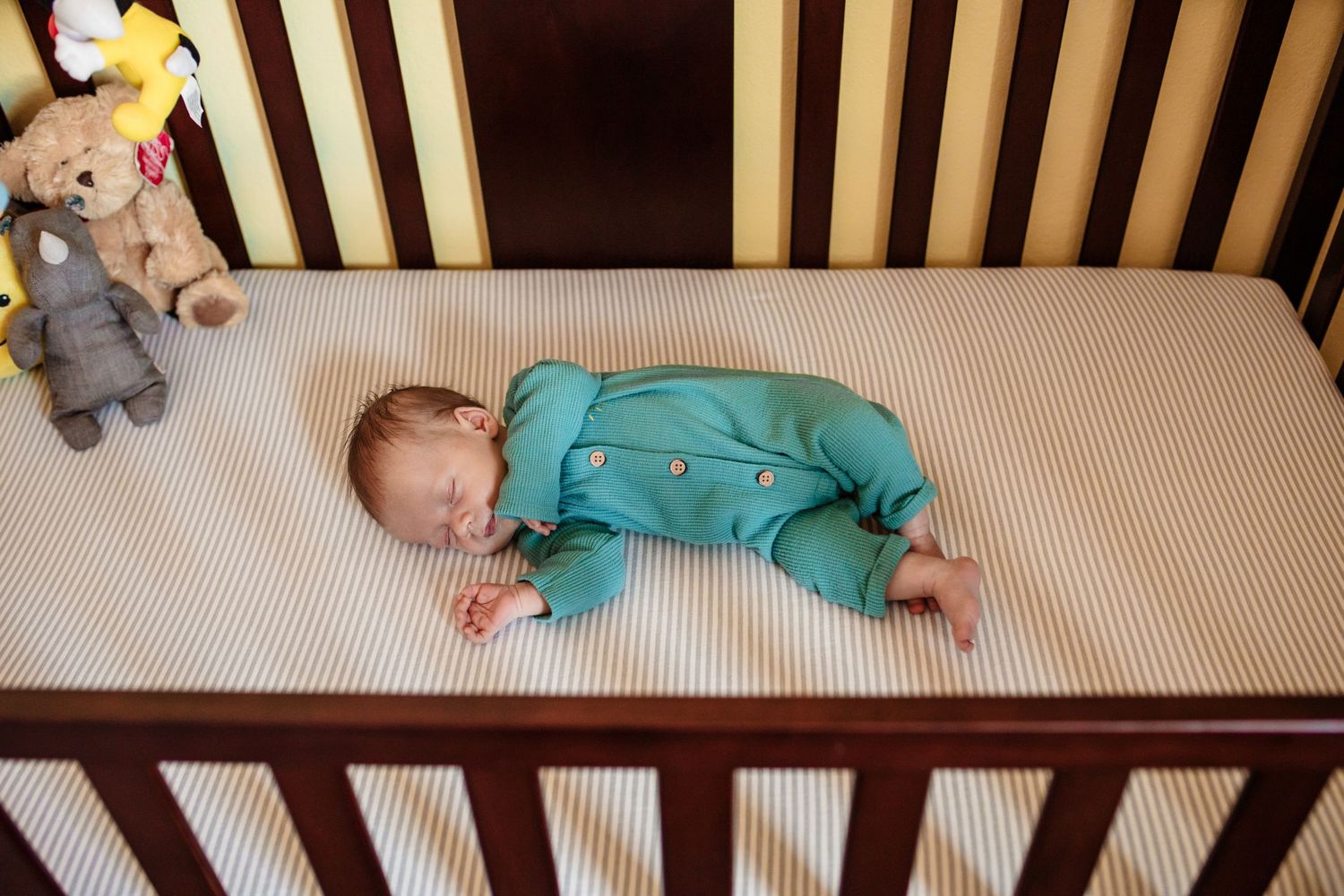 A sleeping baby wearing teal pajamas rests in a wooden crib with stuffed animal toys.