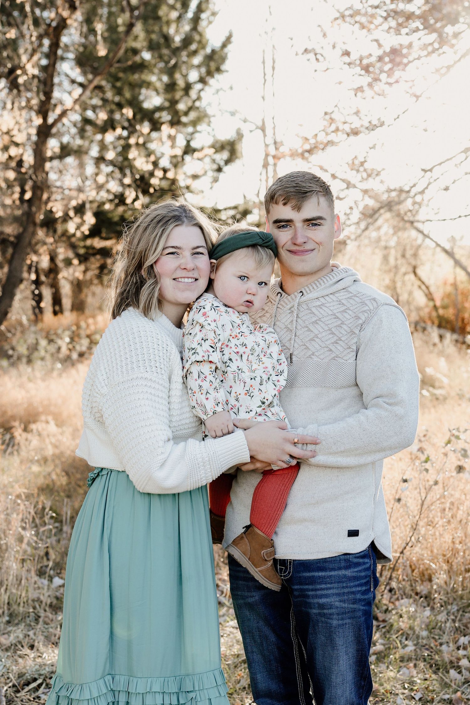 Young family of three smiling outdoors in warm autumn light with trees in the background.