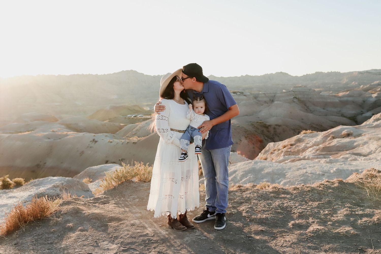Couple sharing kiss while holding baby at golden hour in scenic desert badlands landscape.