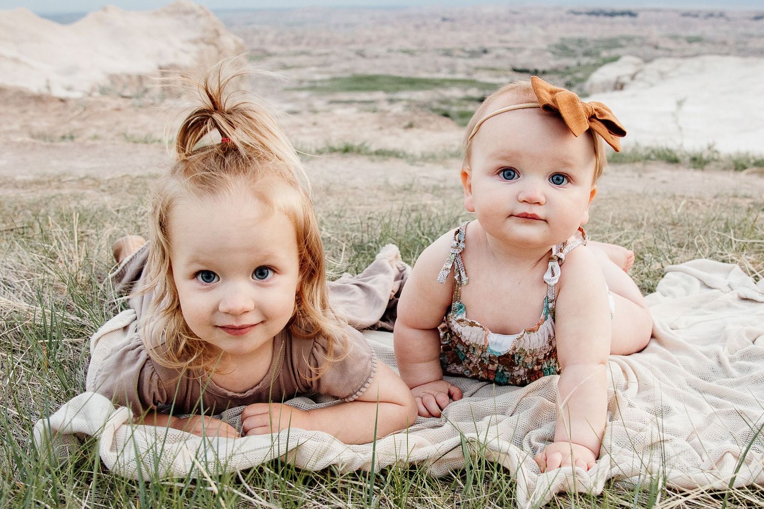 Two adorable baby sisters lying on a blanket in a grassy field with scenic badlands in the background.