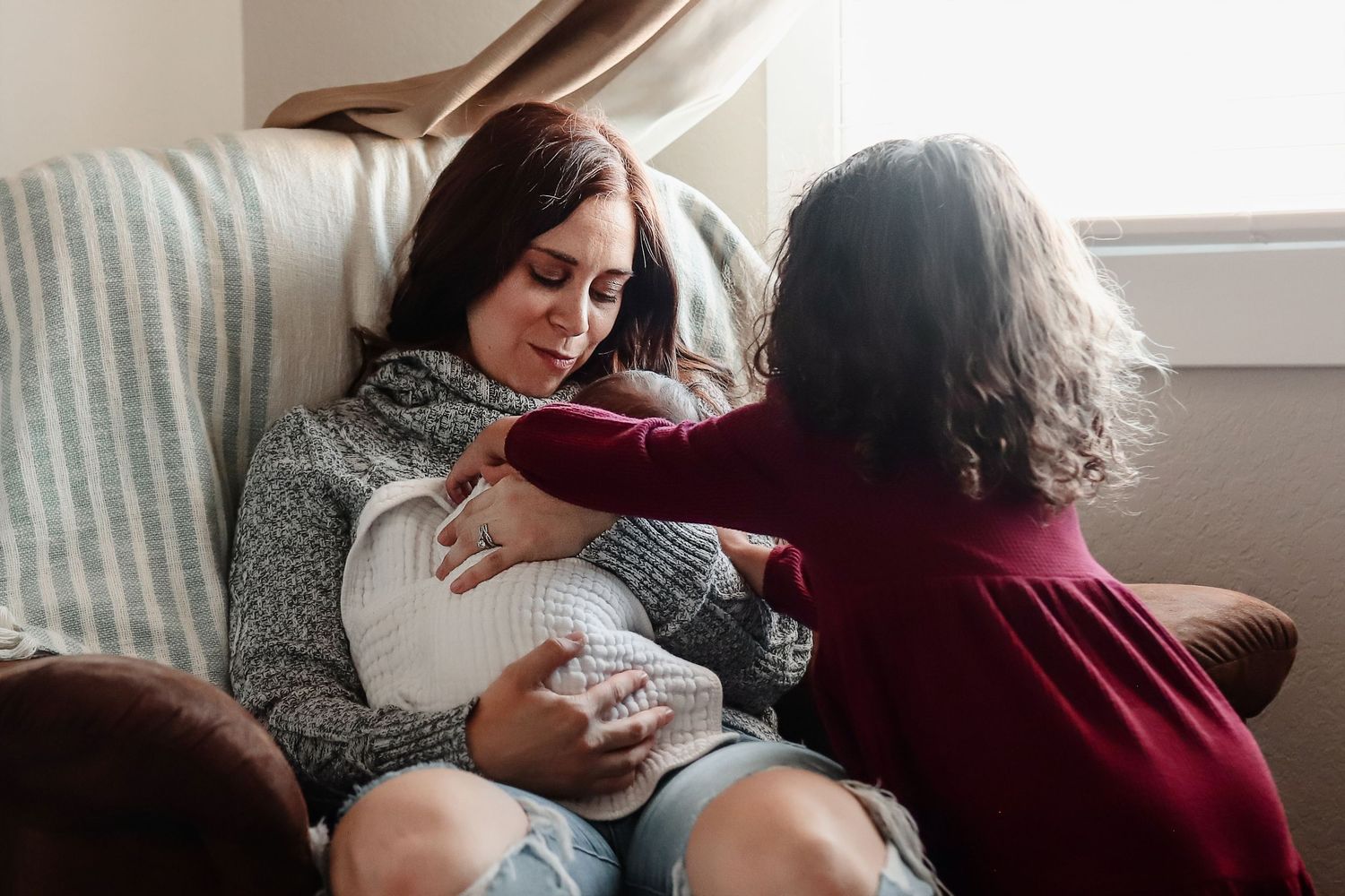 Mother in gray sweater tenderly holds newborn baby while another person assists in a cozy living room chair.