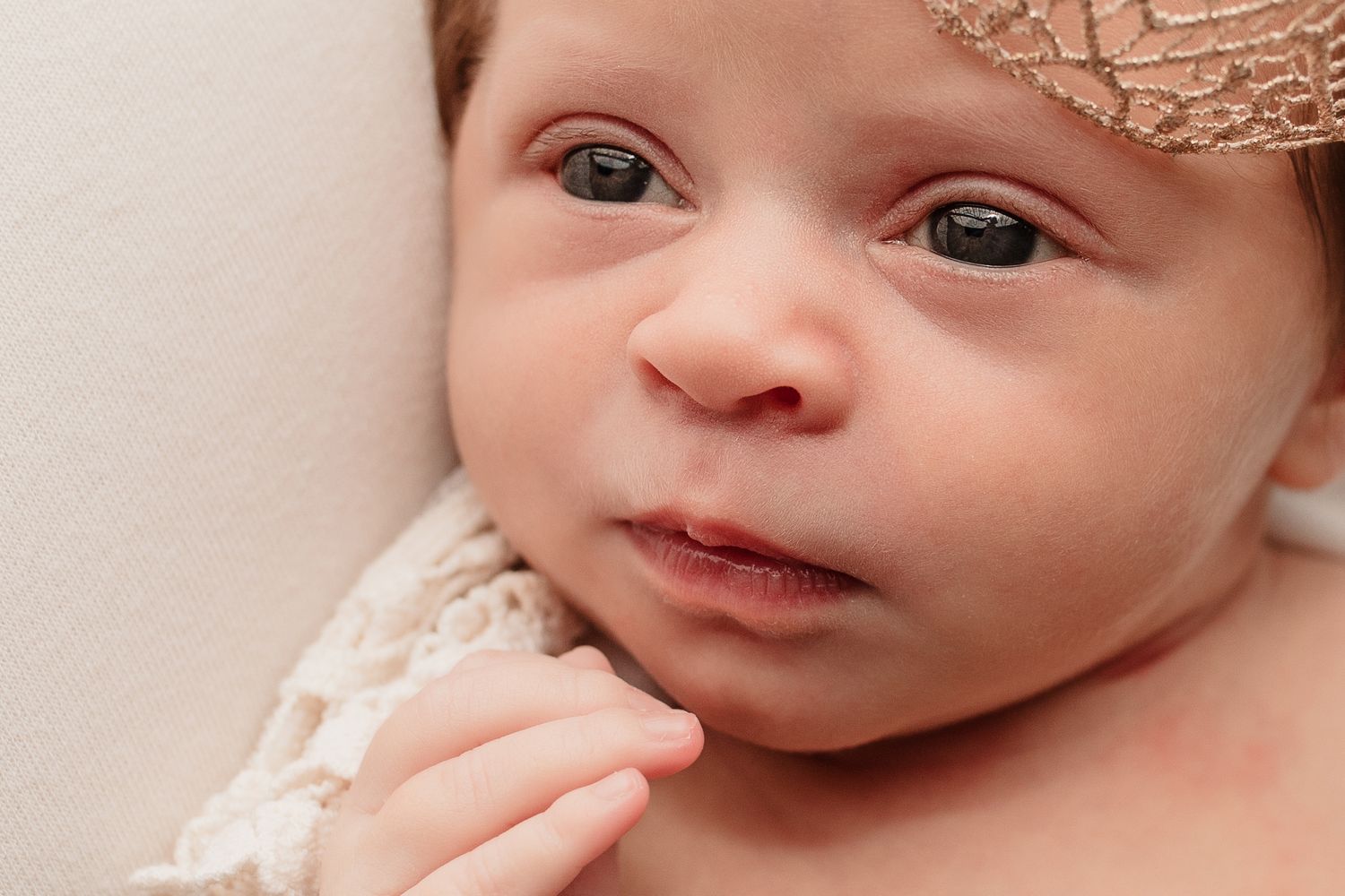 Close-up portrait of an alert newborn baby wearing a delicate lace headband with tiny hand visible.