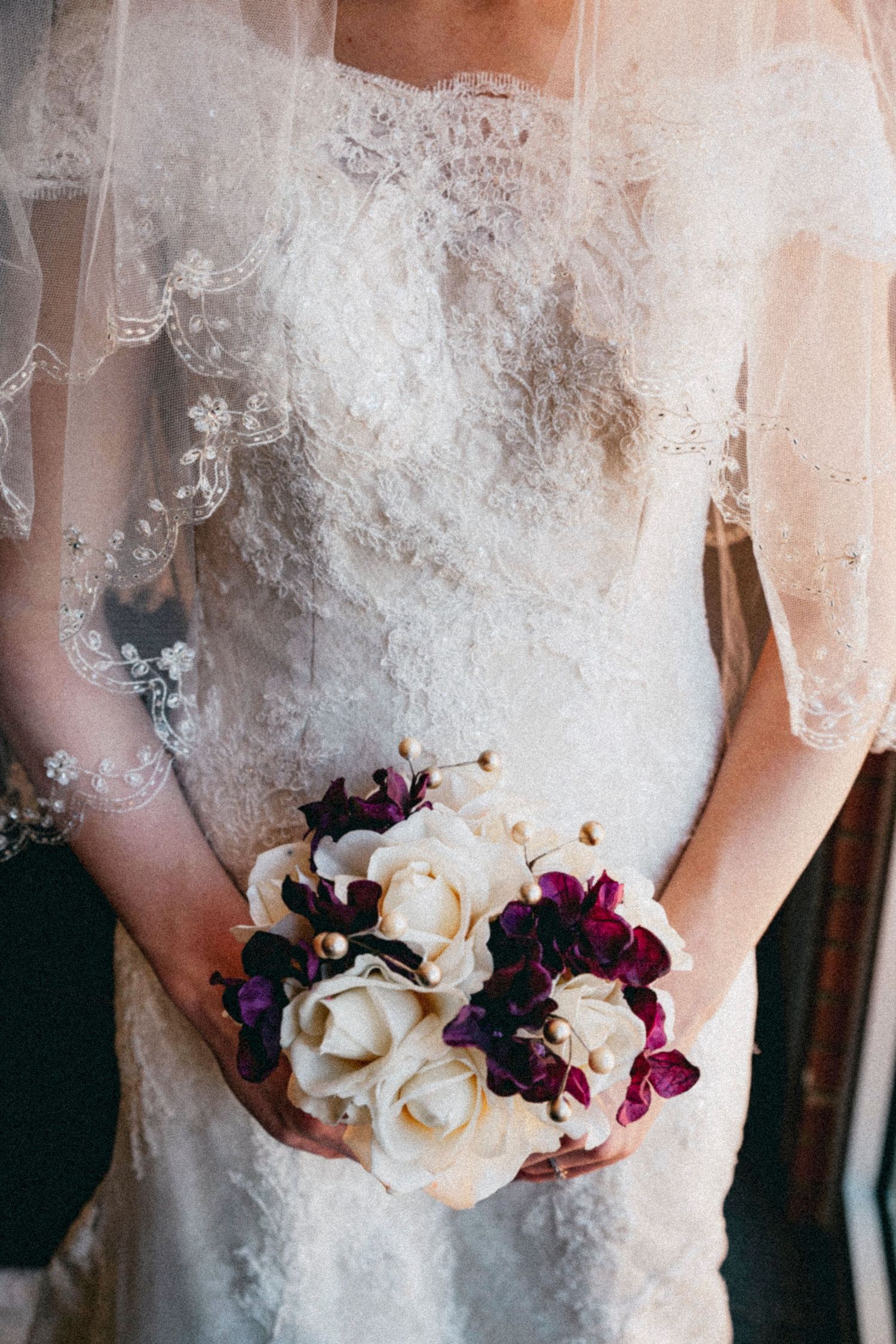 Bride in lace wedding dress and veil holding a bouquet of white roses and purple orchids.