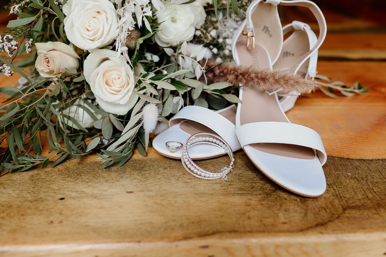 White bridal heels, diamond rings, and a white rose bouquet with greenery arranged on a rustic wooden surface.