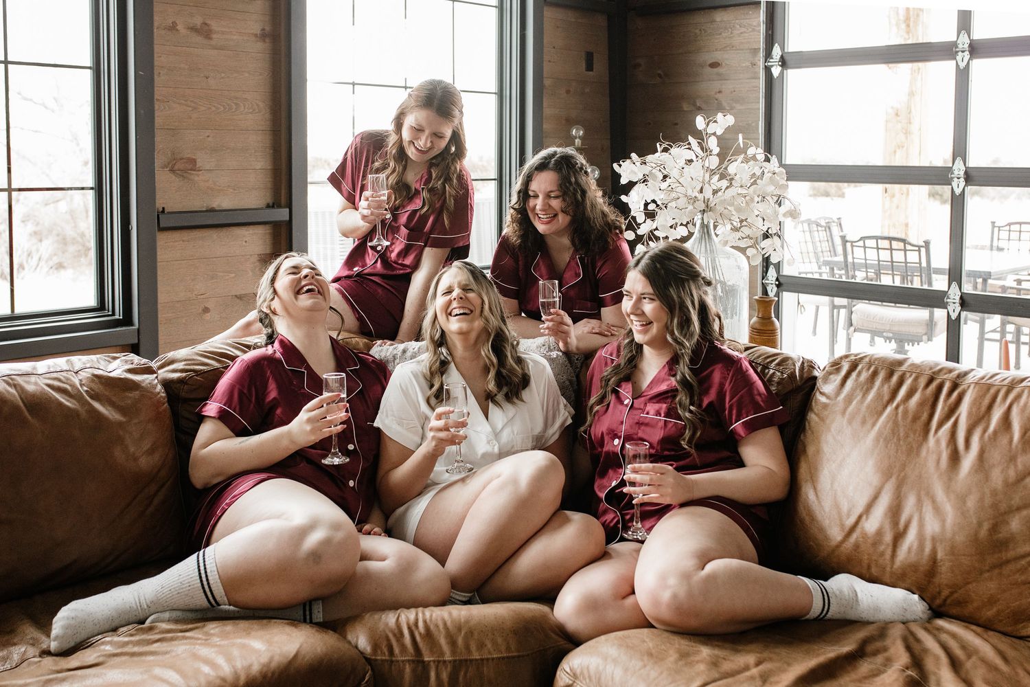 Five bridesmaids in burgundy satin pajamas laughing together on a leather couch during a bridal party getting ready session.