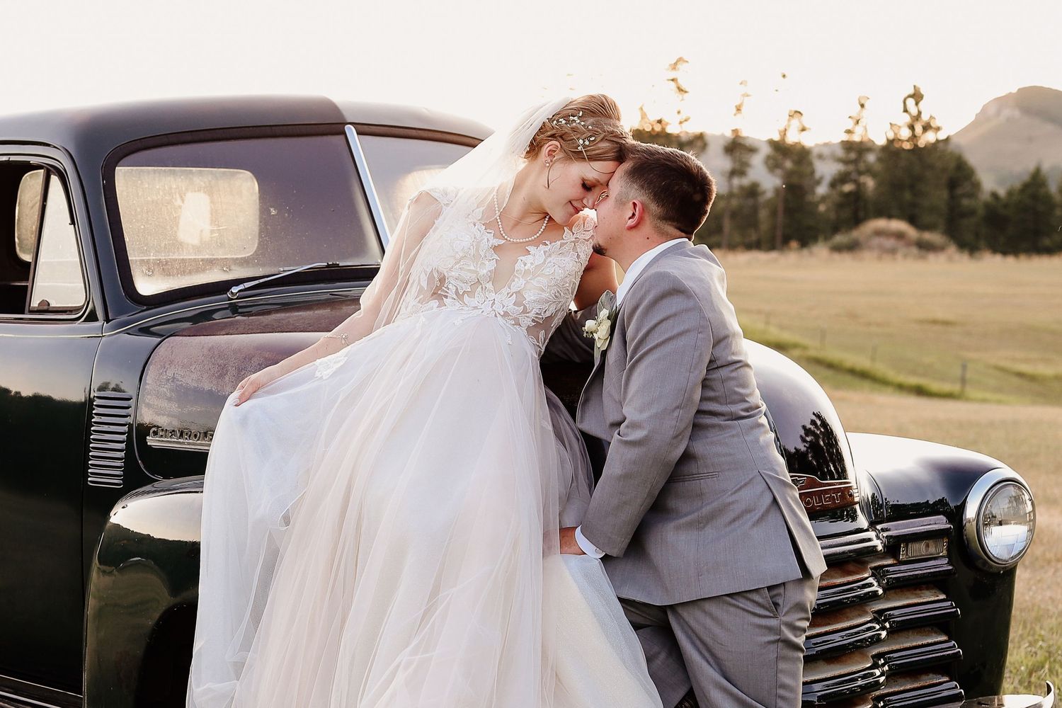 Bride and groom share romantic moment leaning in for a kiss beside a vintage black classic car outdoors.