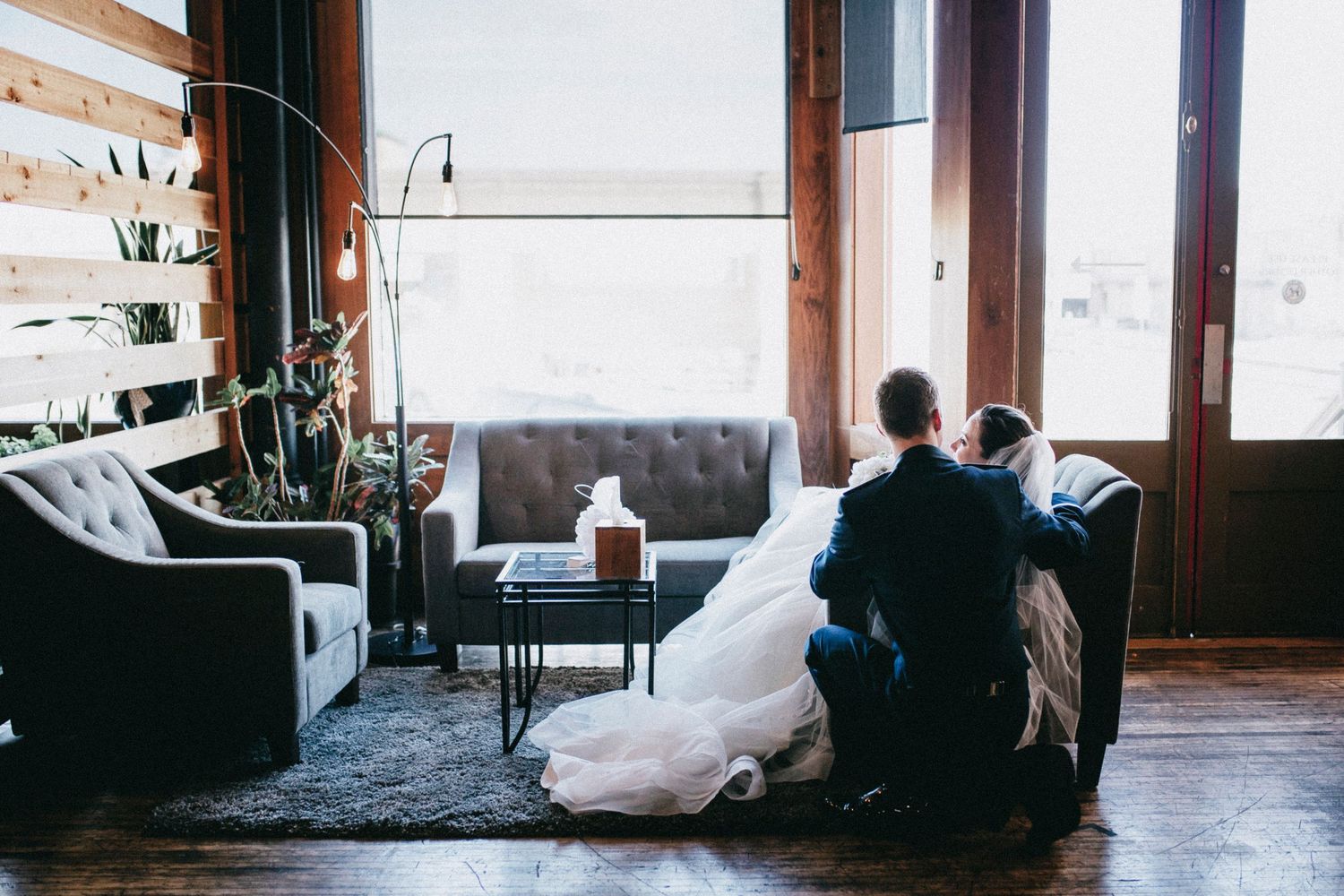 Bride and groom share an intimate moment on a couch by large windows in a rustic wedding venue.