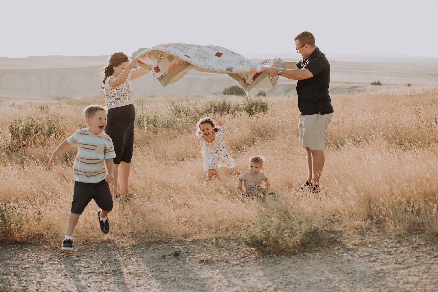 Family playing with a blanket in a golden field, with children running and laughing on a sunny day.