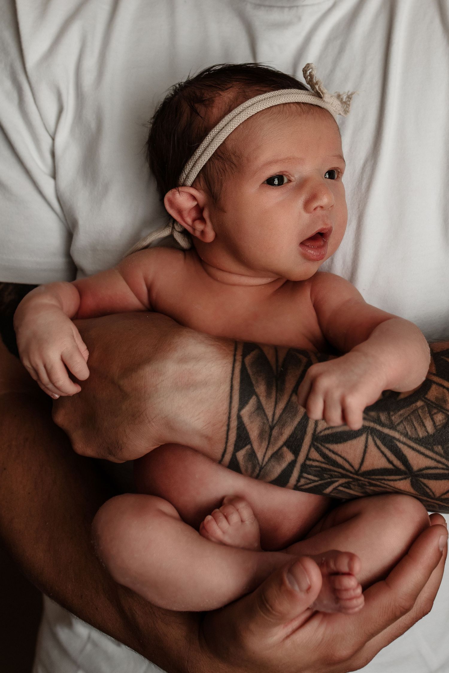 Alert newborn baby girl with headband resting on tattooed fathers hands and chest.