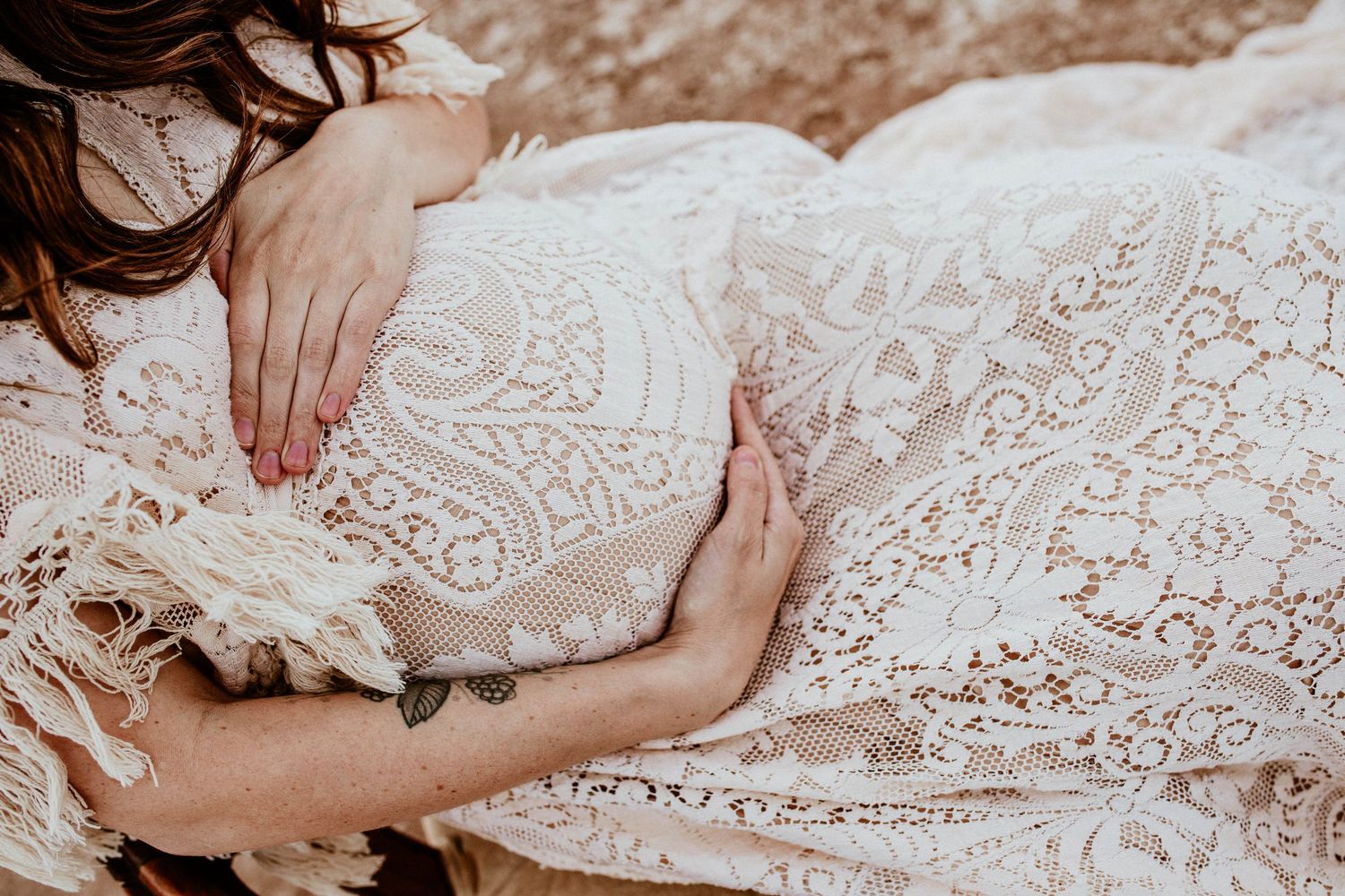 Overhead close-up of pregnant belly in intricate cream lace boho dress with fringe detail.