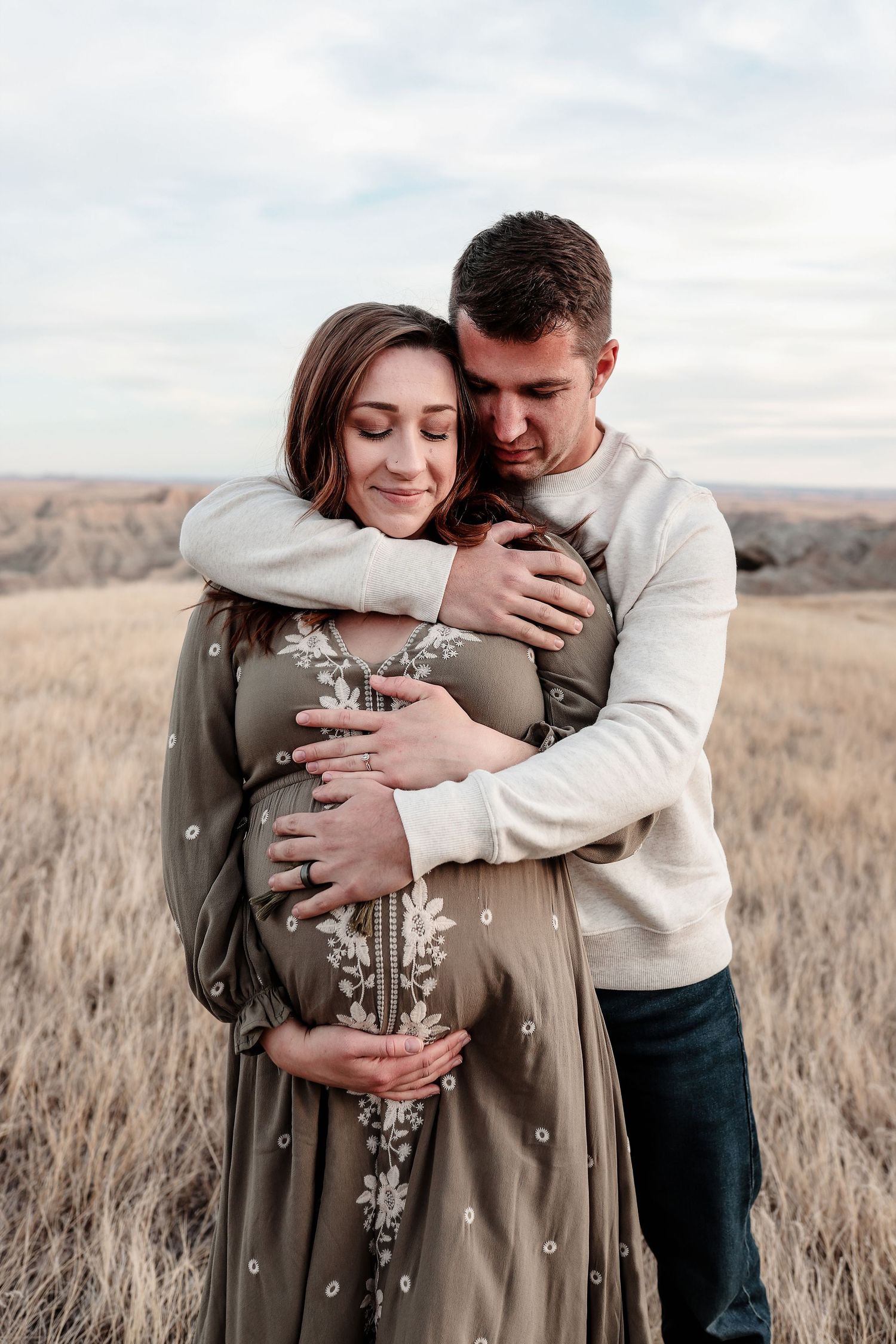 Man tenderly embracing pregnant woman in olive embroidered dress in golden field.
