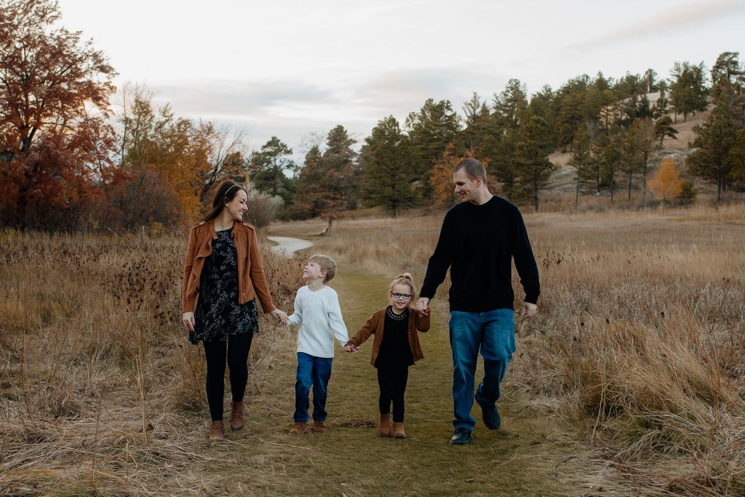 Family of four walking hand in hand through a scenic autumn meadow with colorful fall foliage.