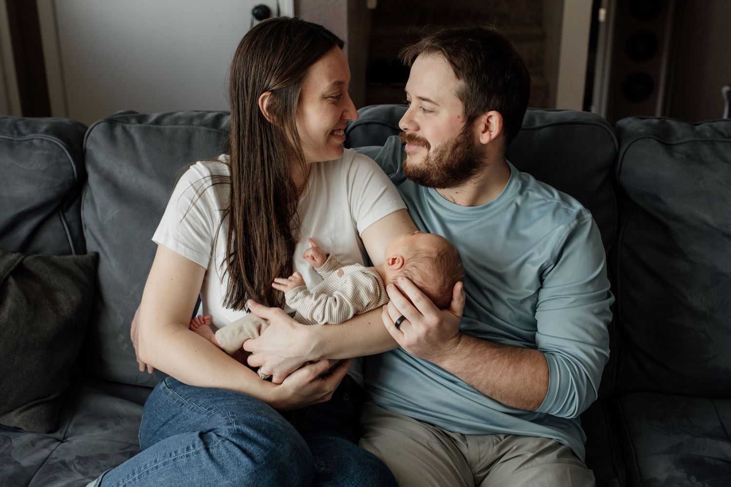 Family shares tender moment while holding newborn baby together on grey couch at home.