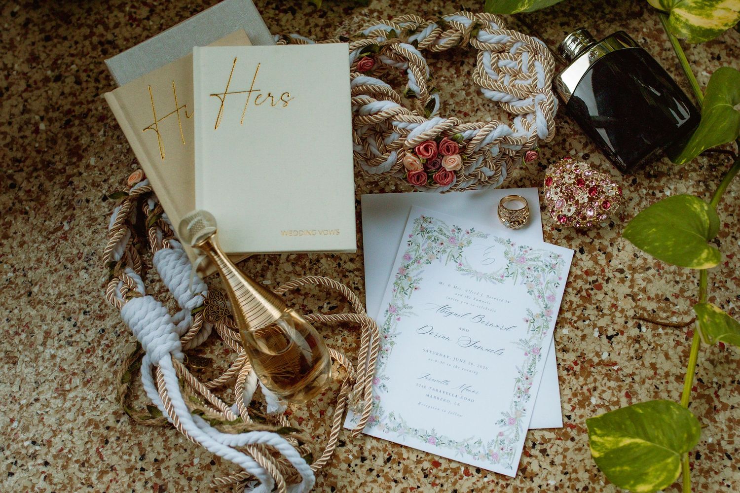 Wedding flat lay featuring invitation suite, unity cord, perfume bottle, and decorative accessories on marble surface.