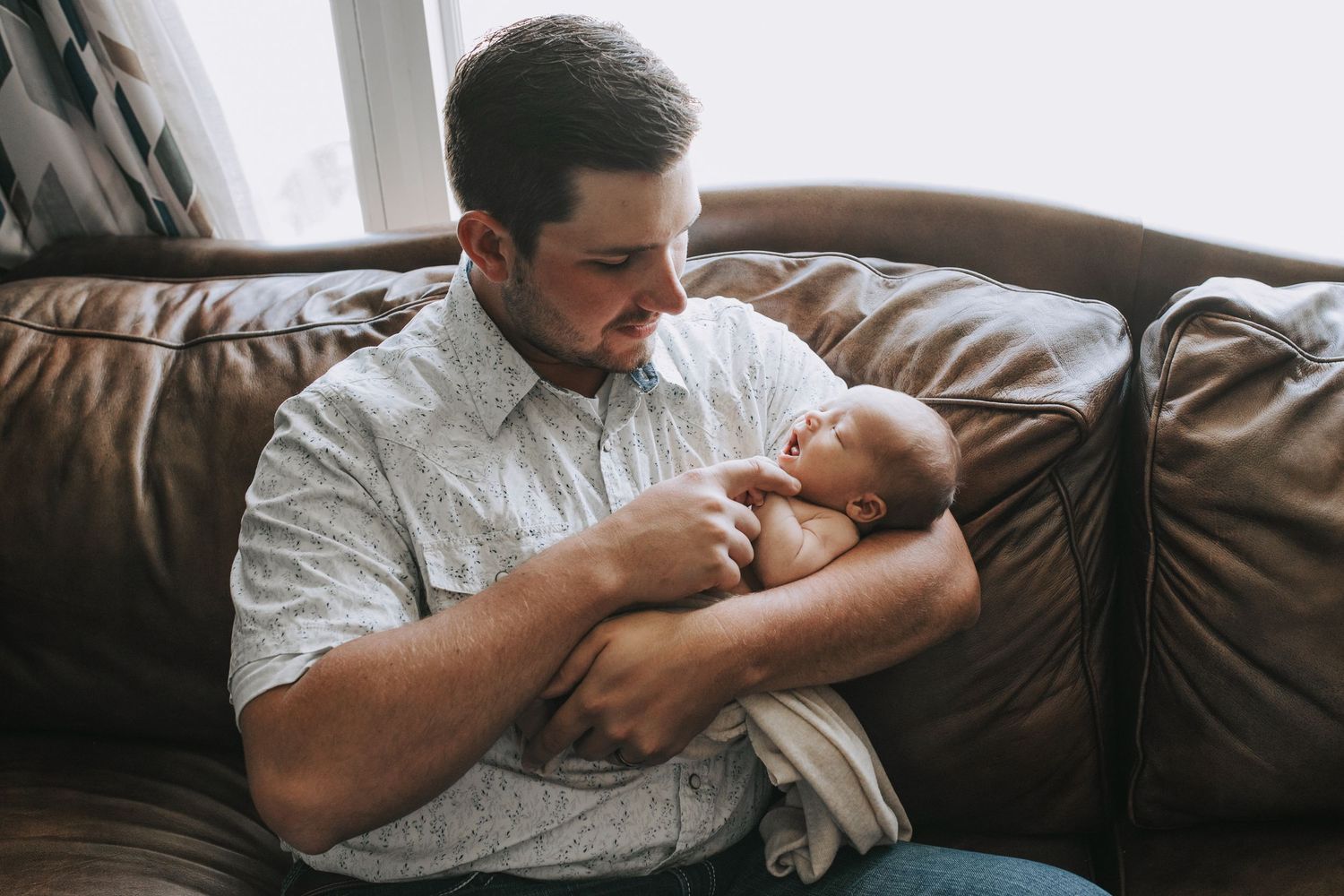 A bearded father tenderly cradles his newborn baby on a brown leather couch near a window.