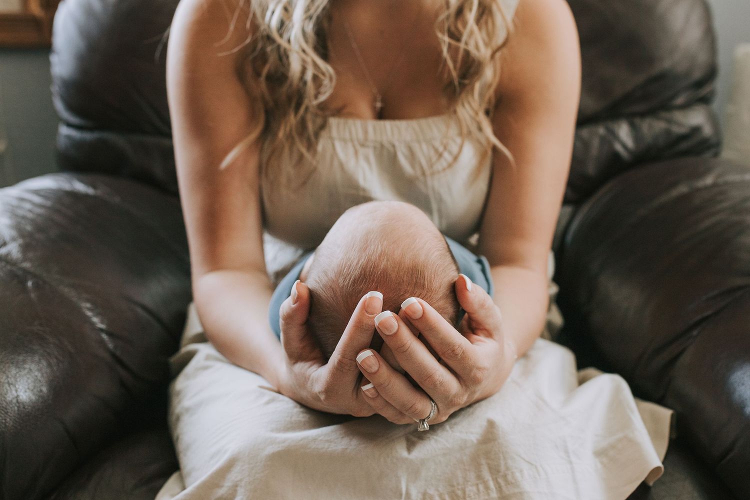 Mother gently cradling newborn baby's head in her hands while seated on a leather sofa.