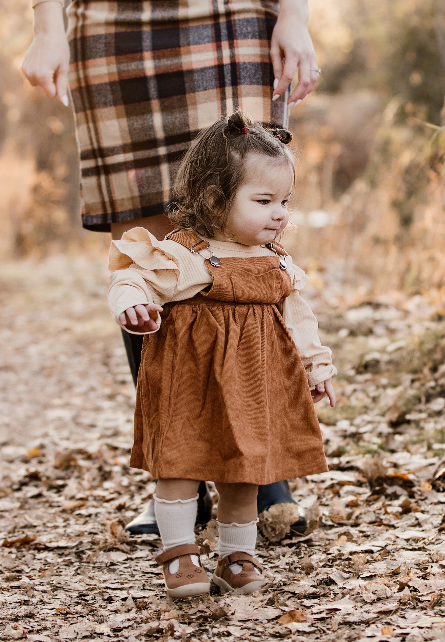 Toddler girl in rust pinafore dress walking on autumn leaves path outdoors.