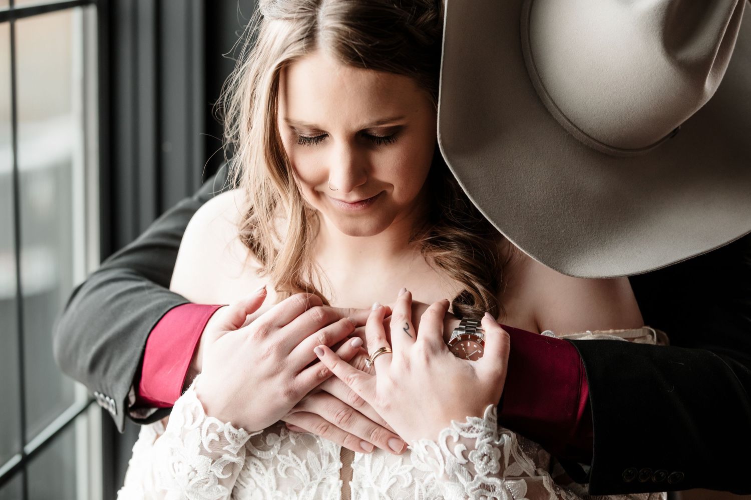 Groom in cowboy hat embracing bride in lace wedding dress, hands clasped together in romantic wedding portrait.