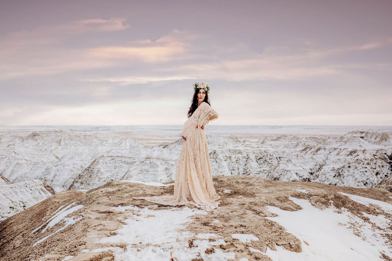 Pregnant woman in flowing cream gown and floral crown stands on snowy badlands ridge at sunset.