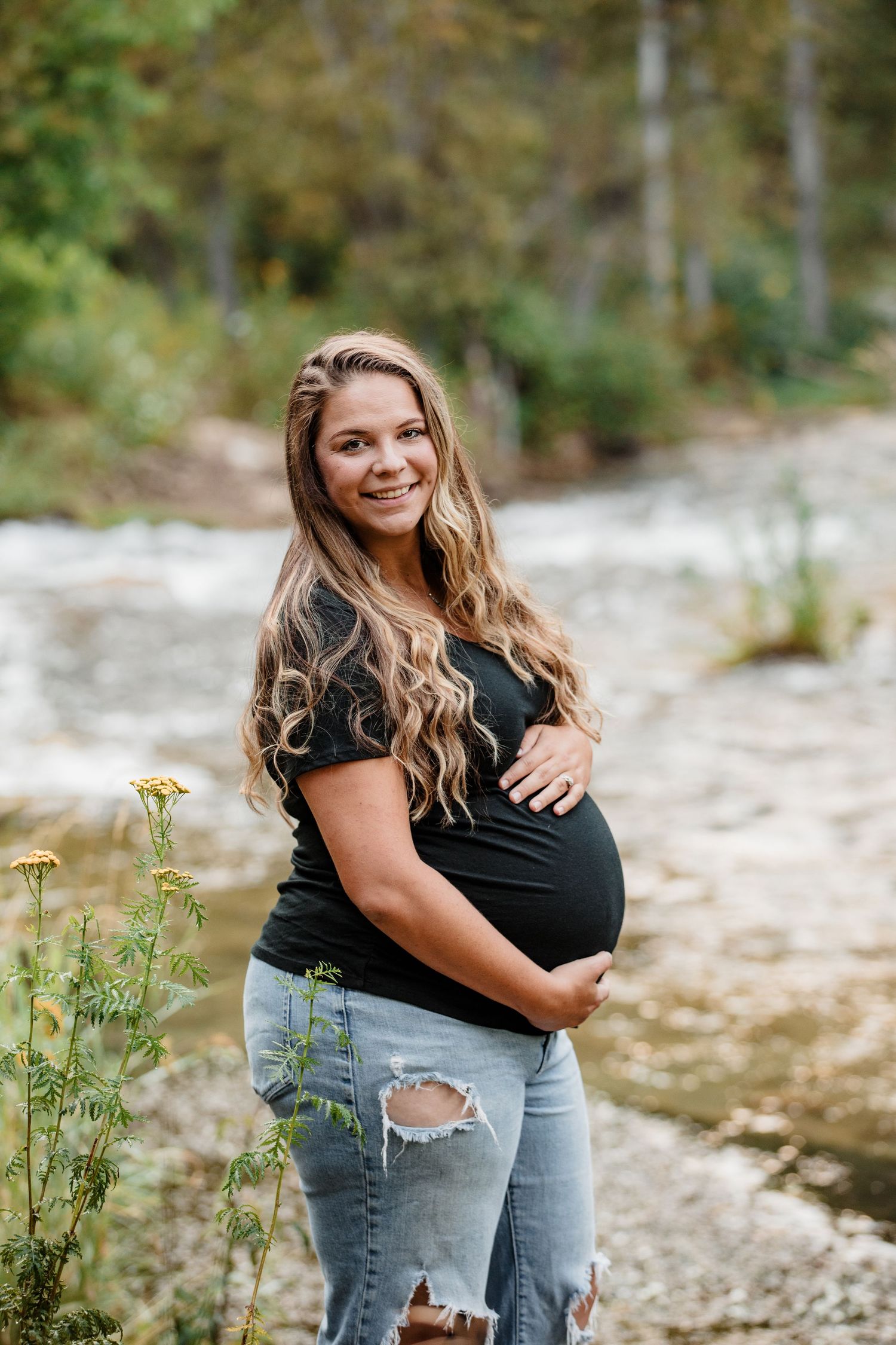 Smiling pregnant woman with long wavy hair in black top and ripped jeans cradling bump near river.