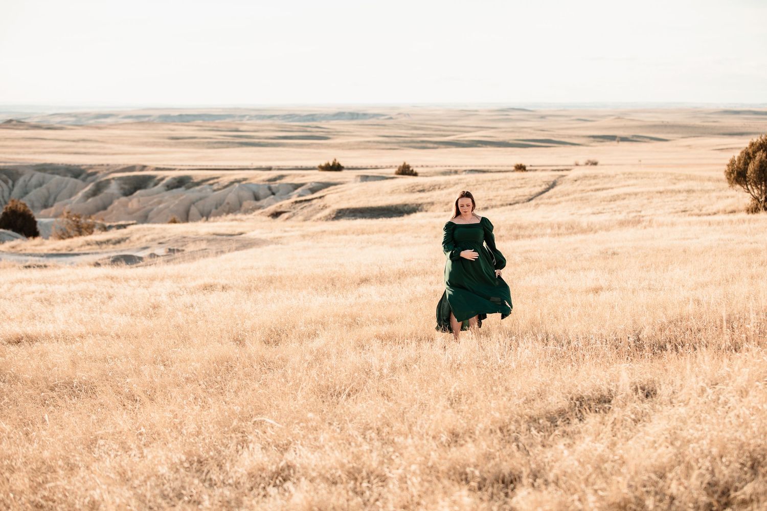 Someone in an emerald green dress stands alone in a vast golden prairie landscape at sunset.