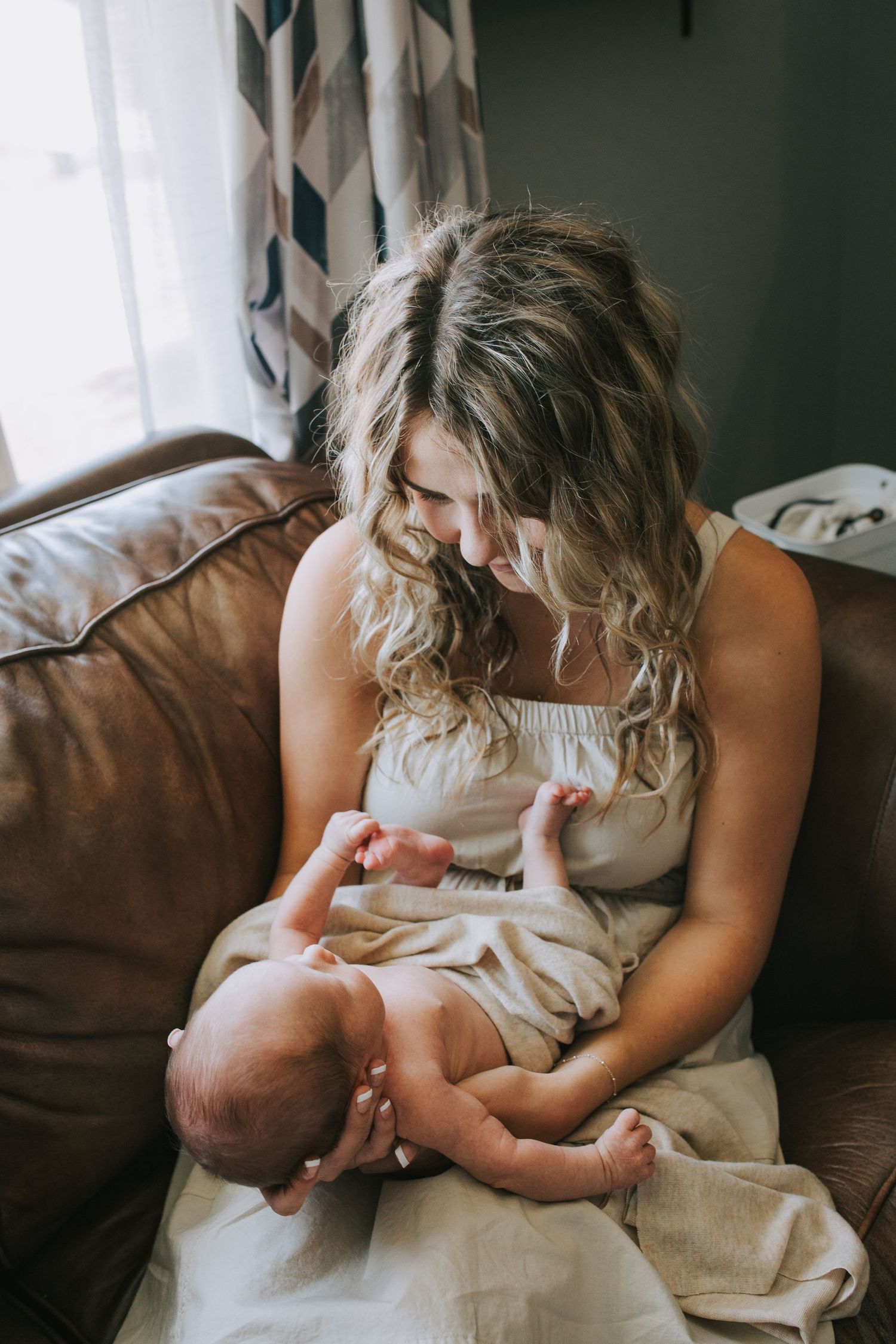 A mother with curly blonde hair smiles down at her newborn baby while seated on a brown leather couch.