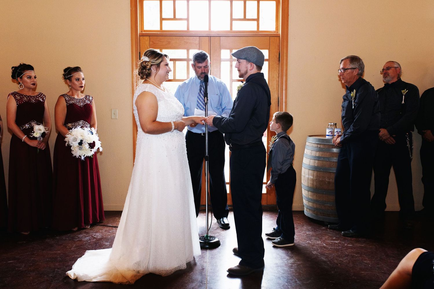 A bride and groom exchange vows at an indoor wedding ceremony with bridesmaids in burgundy and a young ring bearer.