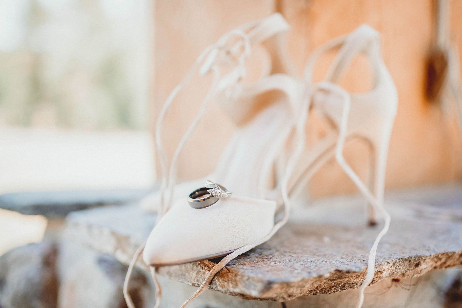 White strappy bridal heels with wedding rings resting on top, displayed on a rustic stone surface.