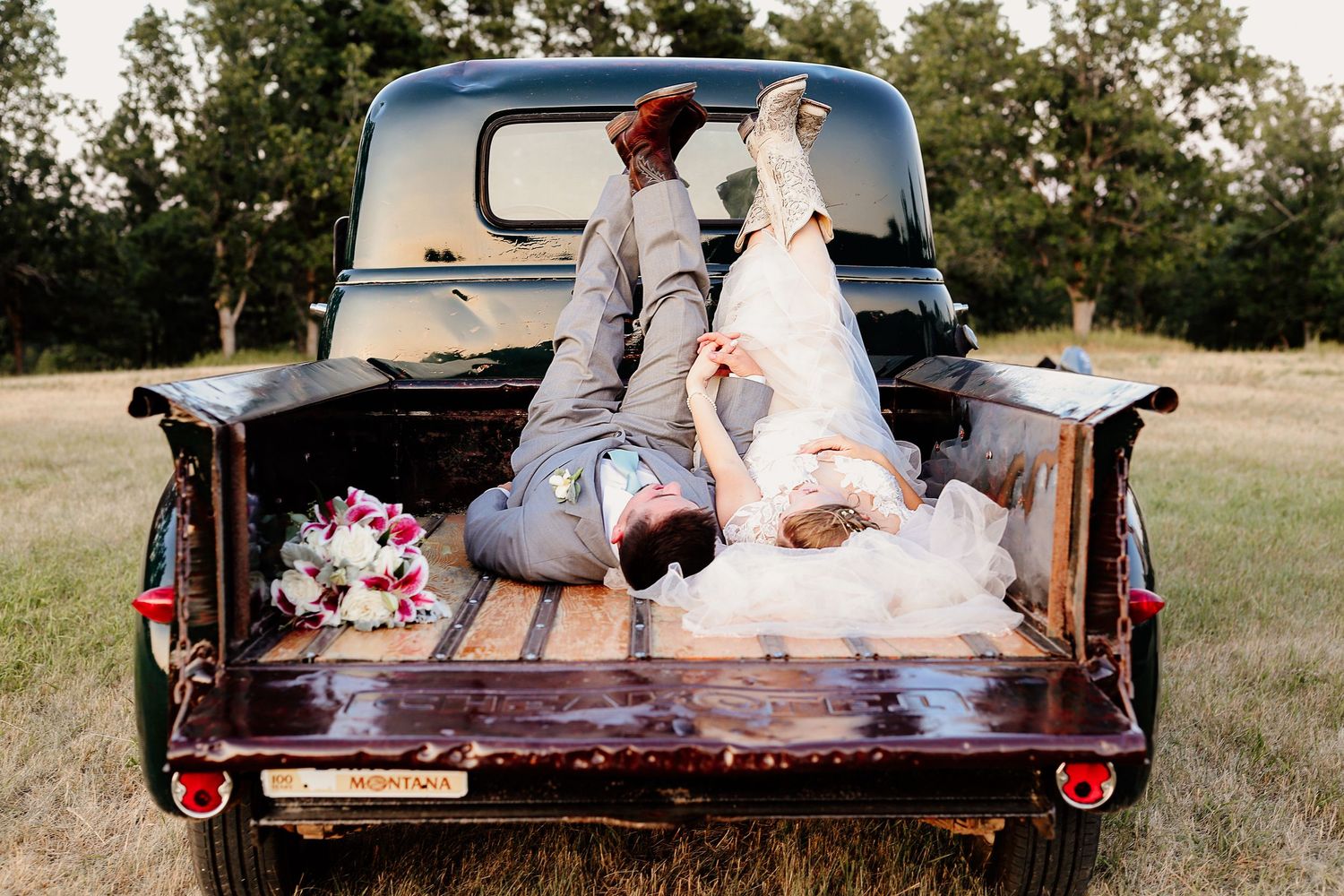 Bride and groom lying in vintage black truck bed with wedding bouquet at rustic outdoor wedding.